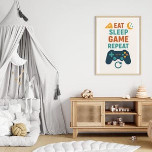 Könnte beinhalten: Ein gerahmter Druck mit dem Text "EAT SLEEP GAME REPEAT" in Orange, Türkis und Grün, mit einer Pizzaschnitte, einem Mond und einer Gamecontroller-Grafik. Der Druck befindet sich in einem hellen Holzrahmen und wird in einem Kinderzimmer ausgestellt.