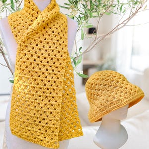 Pode incluir: Um conjunto de cachecol e chapéu bucket de crochê amarelo mostarda. O cachecol, com uma trama aberta, é drapeado sobre um manequim. O chapéu a condizer é exibido numa cabeça de manequim separada. Ambos os artigos são feitos com um padrão de crochê semelhante. O fundo é macio e neutro.