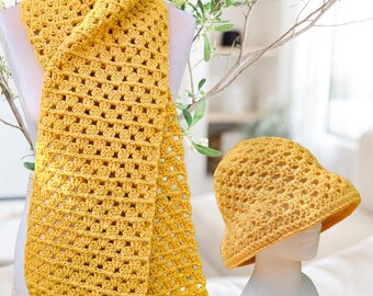 May include: A handmade, mustard yellow crochet scarf and matching bucket hat. The scarf is draped over a mannequin, showcasing its open weave design. The hat sits on a separate mannequin head, also displaying the same crochet pattern. The items are displayed against a soft, neutral background.