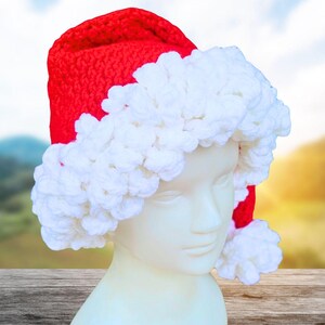 Pode incluir: Um gorro de Pai Natal vermelho de croché com uma barra e um pompom brancos e fofos. O chapéu é exibido numa cabeça de manequim. O fundo está desfocado, com uma superfície de madeira em primeiro plano.