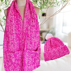 Pode incluir: Uma echarpe de crochê rosa choque com dois bolsos e um gorro a condizer com um pompom rosa e branco. A echarpe é drapeada em um manequim. O gorro e a echarpe são feitos de um fio espesso e texturizado.