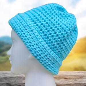 May include: A light blue, handmade crochet beanie hat. The hat is made of a soft, textured yarn and features a folded brim. The hat is displayed on a white mannequin head against a blurred outdoor background.