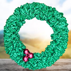 May include: A handmade green knitted wreath with a textured appearance. The wreath is adorned with three small, shiny, pink ornaments. The background is a blurred image of a sky and a wooden surface.