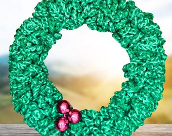 Könnte beinhalten: Ein handgefertigter, grüner Strickkranz mit strukturierter Optik. Der Kranz ist mit drei kleinen, glänzenden, rosa Ornamenten verziert. Der Hintergrund ist ein verschwommenes Bild eines Himmels und einer Holzoberfläche.