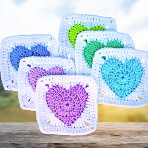 May include: A collection of six handmade crochet squares, each featuring a heart design in various colors. The hearts are in shades of blue, green, and purple, set against a white background. The squares are arranged on a wooden surface.