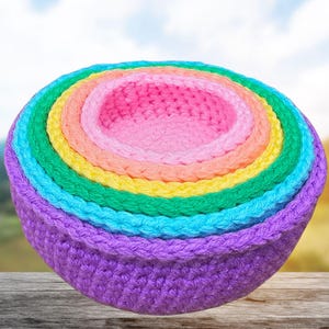 May include: A set of six nested crochet bowls in a rainbow of colors: purple, blue, green, yellow, orange, and pink. The bowls are arranged in a stack, with the smallest bowl at the top. The bowls are made of a soft, textured yarn.