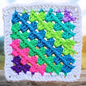 May include: A square, handmade crochet piece featuring a vibrant color palette. The design includes a central pattern of blue, green, pink, and purple, framed by a white border. The texture is clearly visible, showcasing the yarn's individual stitches.