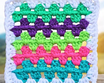 May include: A square, handmade crochet piece with a white border. The interior features rows of vibrant colors: green, purple, lime green, pink, and teal. The crochet pattern creates a textured, geometric design, perfect for a blanket or pillow.