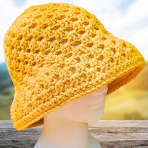 Pode incluir: Um chapéu de balde de crochê amarelo. O chapéu é feito de um fio texturizado de trama aberta e tem uma coroa arredondada e uma aba inclinada para baixo. O chapéu é exibido em uma cabeça de manequim branca contra um fundo desfocado ao ar livre.