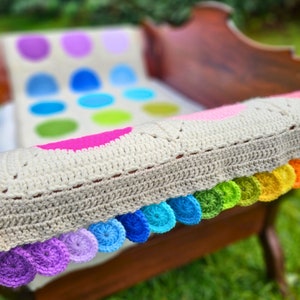 Op de afbeelding: Een gehaakt babydekentje met een kleurrijke regenboogrand. Het dekentje is gemaakt van wit garen en heeft een patroon van kleurrijke cirkels.