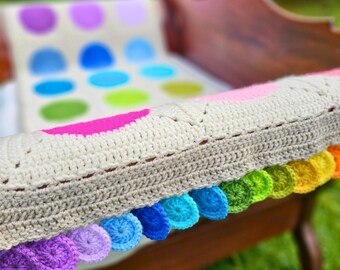 Könnte beinhalten: Eine gehäkelte Babydecke mit einem bunten Regenbogenrand. Die Decke ist aus weißem Garn gefertigt und hat ein Muster aus bunten Kreisen.