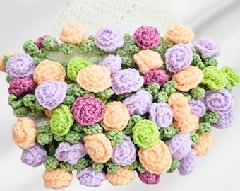May include: A crocheted flower garland with a variety of pastel colors, including pink, purple, peach, and green. The garland is made of small crocheted flowers and leaves.