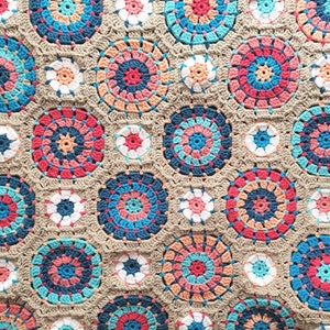 Pode incluir: Uma manta de crochê com um padrão repetitivo de círculos coloridos. Os círculos são feitos de diferentes tons de azul, rosa e laranja. O fundo da manta é de cor marrom claro.