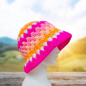 Pode incluir: Um chapéu de balde de crochê colorido em tons de rosa, laranja e branco. O chapéu é exibido em uma cabeça de manequim branca. A aba é de um fúcsia vibrante. O chapéu é feito de um fio macio e texturizado.