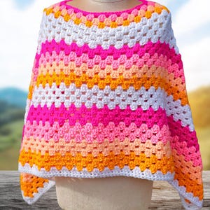 May include: A colorful, handmade crochet poncho in a granny square pattern. The poncho features horizontal stripes of white, bright pink, peach, and orange. The garment is displayed on a mannequin, showcasing its open, flowing design.