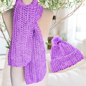 Pode incluir: Um conjunto de cachecol e gorro de crochê feitos à mão em roxo vibrante. O cachecol é longo e retangular, drapeado sobre um manequim. O gorro apresenta um pompom a condizer. O conjunto é feito de fio espesso e texturizado.