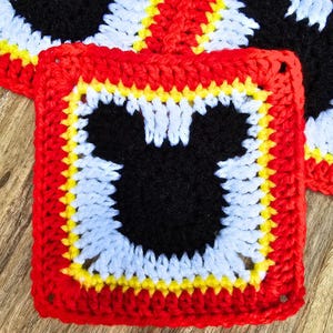 May include: Crocheted square featuring a black Mickey Mouse silhouette on a light blue background, framed by yellow and red borders. The handmade coaster is part of a set, with others visible in the background.