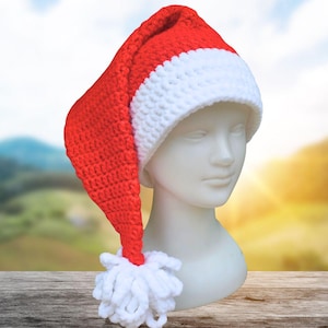 Pode incluir: Um gorro de Natal vermelho e branco de crochê, com formato longo e pontudo e um pompom branco. O chapéu é exibido em uma cabeça de manequim branca, em um fundo desfocado.