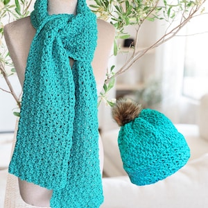 Pode incluir: Um conjunto de cachecol e gorro de crochê turquesa. O cachecol é longo e retangular, com um padrão de ponto texturizado. O gorro tem uma cor turquesa combinando e um pompom de pele sintética marrom. O conjunto é exibido em um manequim.