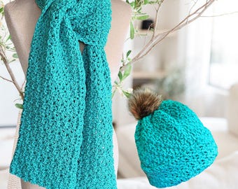 May include: A turquoise crochet scarf draped on a mannequin, paired with a matching beanie featuring a brown faux fur pom-pom. The scarf is long and rectangular, with a textured stitch pattern. The beanie is a close-fitting style.