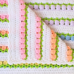 May include: A white crocheted baby blanket with a colourful striped pattern. The blanket features a scalloped edge and is made with a soft, textured yarn.