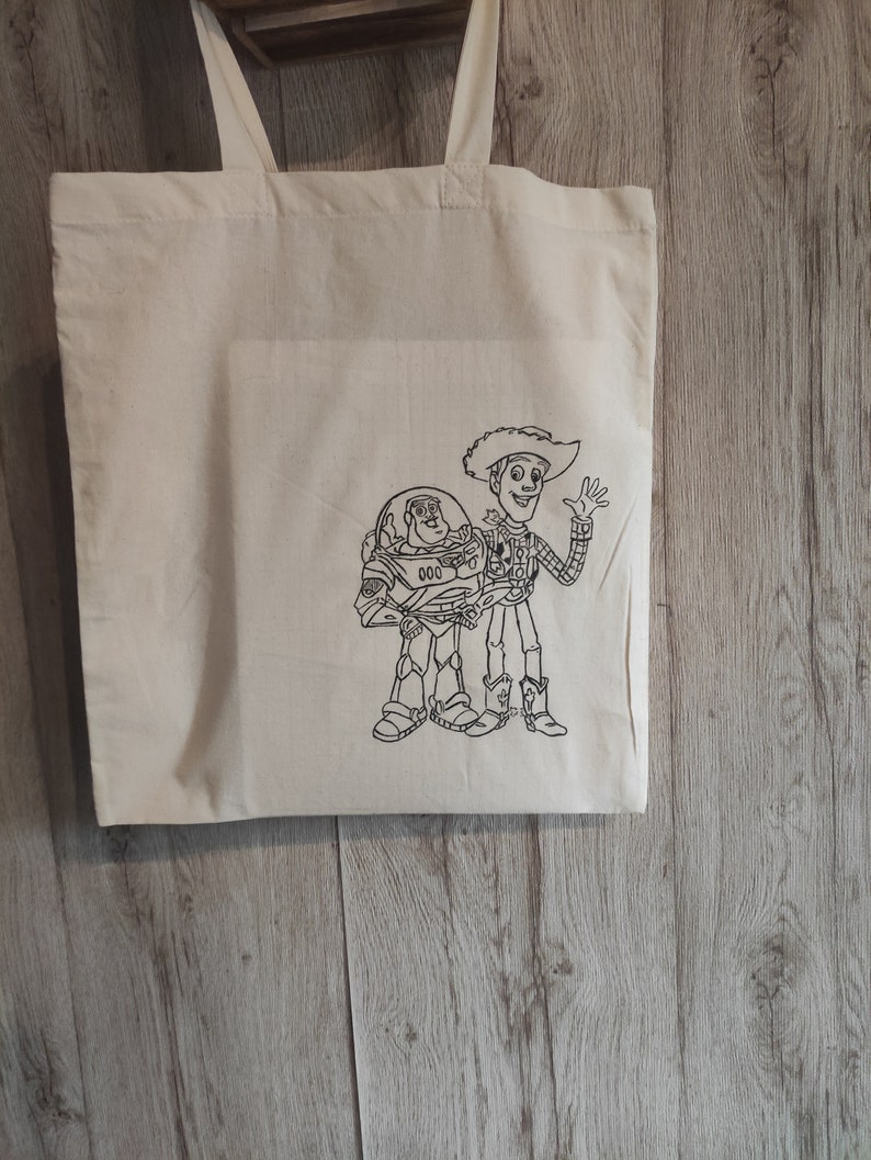 Puede incluir: Una bolsa de lona blanca con un dibujo de l&iacute;nea negra de Woody y Buzz Lightyear de Toy Story. Los personajes est&aacute;n uno al lado del otro, con Woody saludando.
