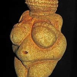 Puede incluir: Una escultura de piedra de color amarillo p&aacute;lido, tallada a mano, que representa una figura femenina con rasgos exagerados. La escultura es estilizada y abstracta, con un cuerpo redondeado y lleno y una cabeza grande con una superficie texturizada.