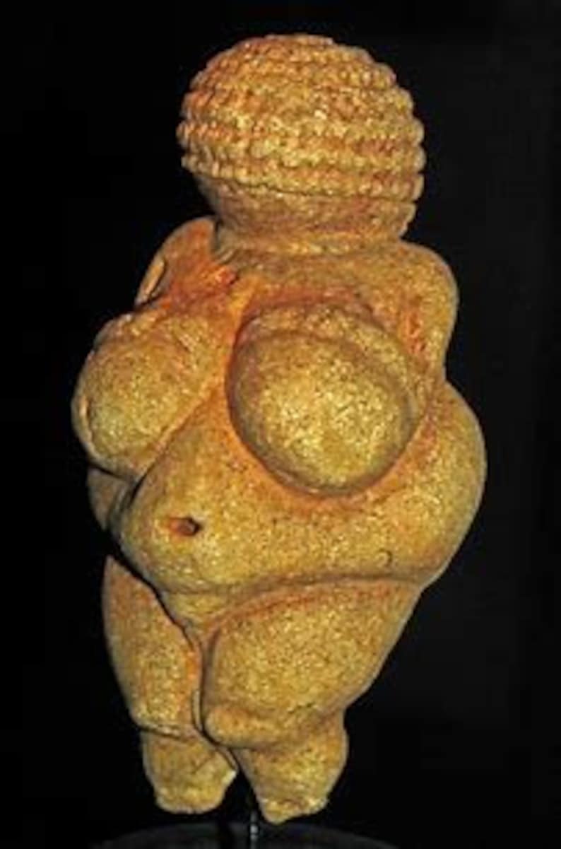 Puede incluir: Una escultura de piedra de color amarillo p&aacute;lido, tallada a mano, que representa una figura femenina con rasgos exagerados. La escultura es estilizada y abstracta, con un cuerpo redondeado y lleno y una cabeza grande con una superficie texturizada.