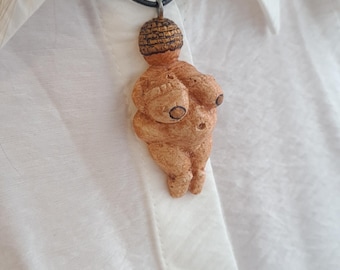 Puede incluir: Un collar con un colgante de cerámica marrón que representa una figura femenina estilizada. El colgante tiene una superficie texturizada y se lleva en un cordón negro.