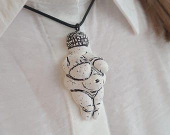 Puede incluir: Un collar con un colgante de cerámica blanca que representa una figura femenina estilizada con detalles en línea negra. El colgante se lleva en un cordón negro.