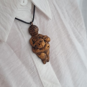 Puede incluir: Un collar con un colgante en forma de figura femenina estilizada de color dorado, con una cabeza redonda y los brazos cruzados sobre el pecho. El colgante est&aacute; colgado de un cord&oacute;n negro.