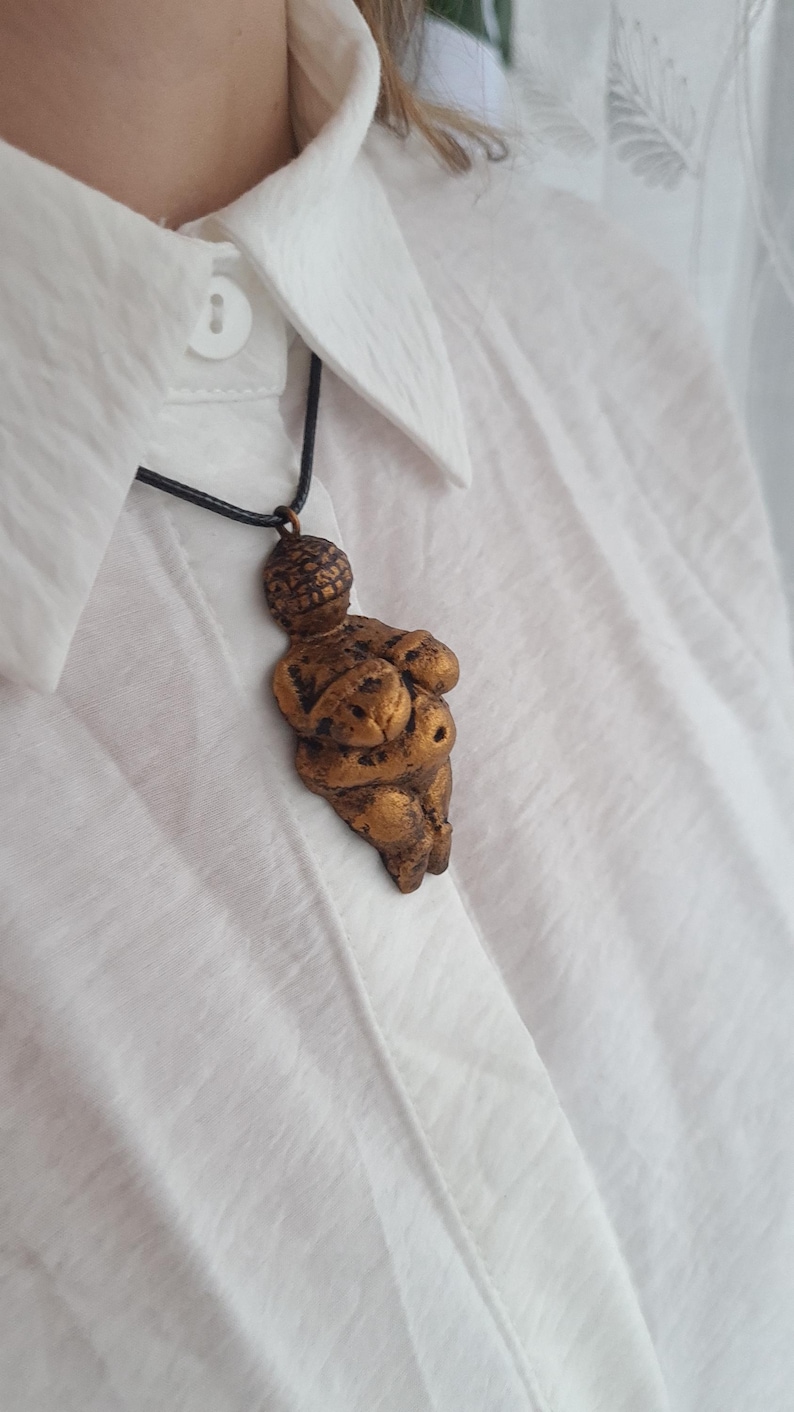 Puede incluir: Un collar con un colgante en forma de figura femenina estilizada de color dorado, con una cabeza redonda y los brazos cruzados sobre el pecho. El colgante est&aacute; colgado de un cord&oacute;n negro.