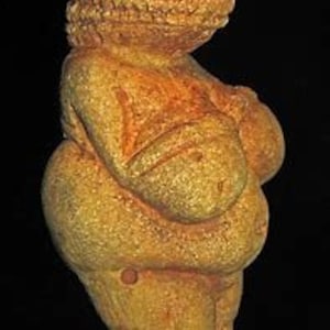 Puede incluir: Una figurita femenina estilizada de color amarillo p&aacute;lido con rasgos exagerados, incluyendo grandes pechos y caderas. La figurita est&aacute; hecha de piedra y tiene una superficie lisa y redondeada.