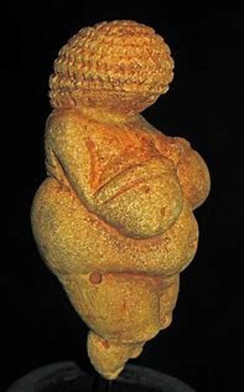Puede incluir: Una figurita femenina estilizada de color amarillo p&aacute;lido con rasgos exagerados, incluyendo grandes pechos y caderas. La figurita est&aacute; hecha de piedra y tiene una superficie lisa y redondeada.