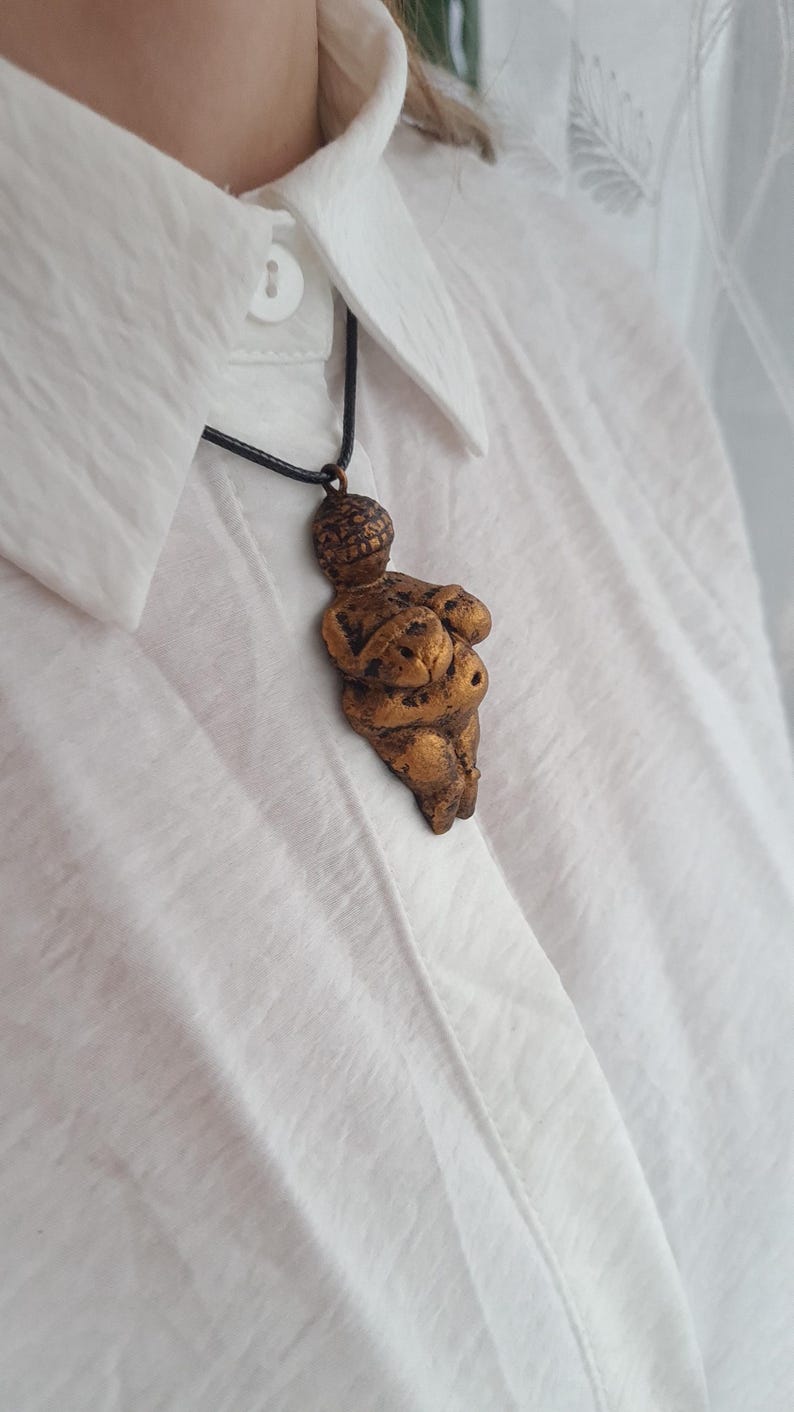 Puede incluir: Un collar de color dorado con un colgante con forma de cuerpo de mujer. El colgante est&aacute; hecho de un material texturizado y tiene una p&aacute;tina marr&oacute;n oscura.