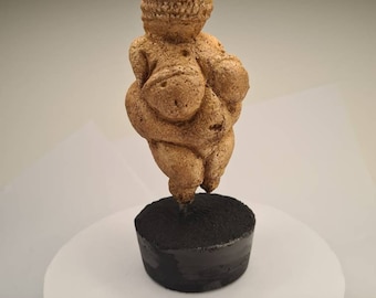 Peut inclure: Une petite sculpture en céramique brune représentant une figure féminine aux traits exagérés. La sculpture est posée sur une base noire.