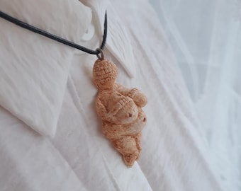 Puede incluir: Un collar con un colgante de cerámica marrón que representa una figura femenina estilizada con vientre redondeado. El colgante cuelga de un cordón negro.