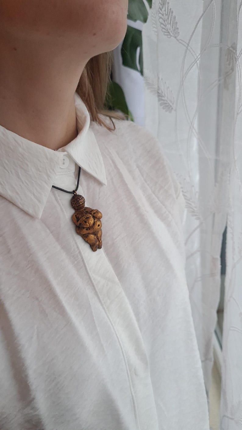 Puede incluir: Un collar dorado con un colgante con forma de cuerpo de mujer. El colgante est&aacute; detallado con una superficie texturizada y una peque&ntilde;a cabeza.