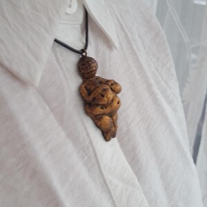 Puede incluir: Un collar con un colgante de color dorado que representa una figura femenina estilizada con la cabeza redonda y los brazos cruzados sobre el pecho. El colgante cuelga de un cord&oacute;n negro.