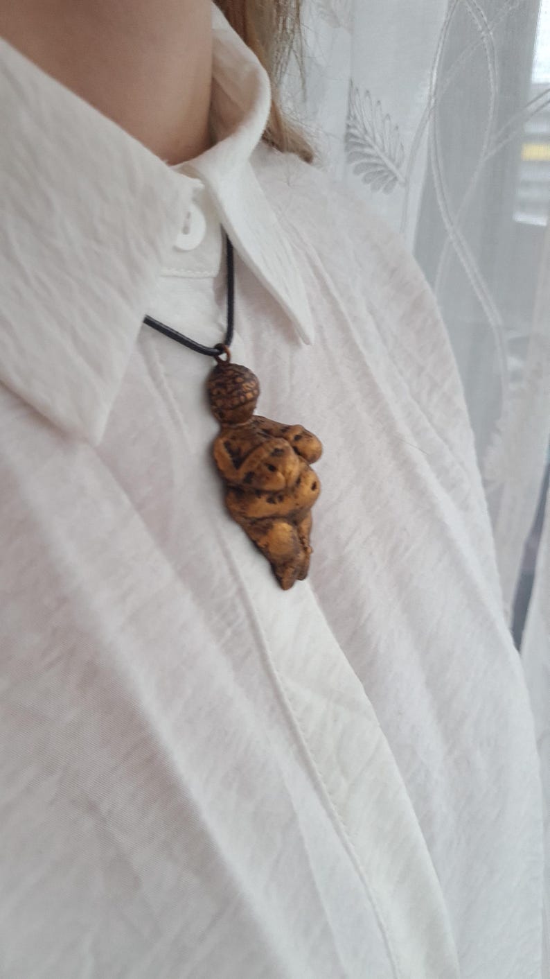 Puede incluir: Un collar con un colgante de color dorado que representa una figura femenina estilizada con la cabeza redonda y los brazos cruzados sobre el pecho. El colgante cuelga de un cord&oacute;n negro.