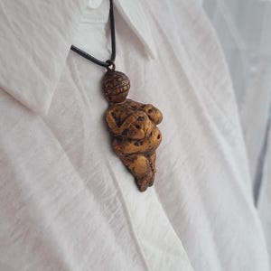Puede incluir: Un collar de color dorado con un colgante con forma de cuerpo de mujer. El colgante est&aacute; hecho de un material texturizado y tiene un dise&ntilde;o ligeramente abstracto.