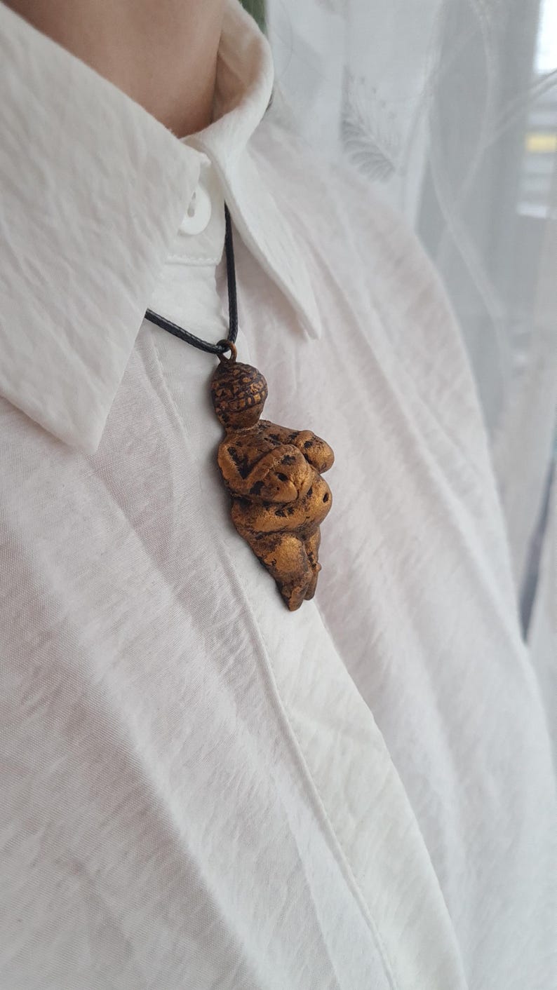 Puede incluir: Un collar de color dorado con un colgante con forma de cuerpo de mujer. El colgante est&aacute; hecho de un material texturizado y tiene un dise&ntilde;o ligeramente abstracto.
