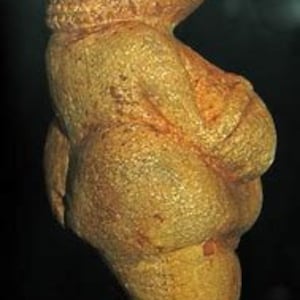 Puede incluir: Una peque&ntilde;a escultura de piedra de color ocre, tallada a mano, que representa una figura femenina. La figura est&aacute; representada con pechos y caderas exagerados, y una cabeza redonda con un peinado estilizado.