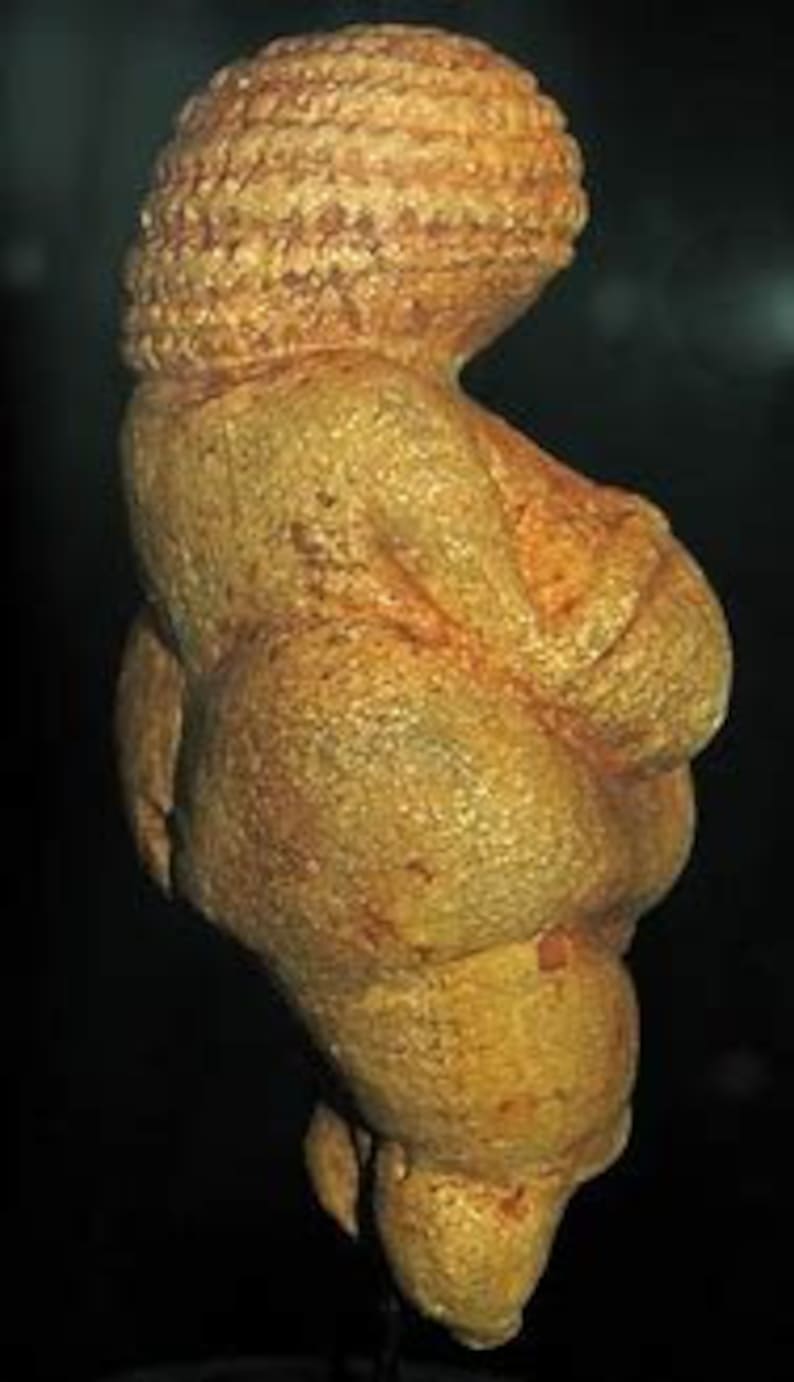 Puede incluir: Una peque&ntilde;a escultura de piedra de color ocre, tallada a mano, que representa una figura femenina. La figura est&aacute; representada con pechos y caderas exagerados, y una cabeza redonda con un peinado estilizado.