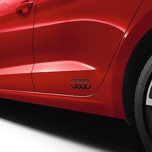 May include: A section of a red car with a black Audi emblem on the side panel. The car's door handle and a portion of the tire are visible. The emblem consists of four interlocking rings.