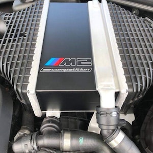 Puede incluir: Primer plano de un compartimento del motor negro con una toma de aire plateada y negra. La toma de aire tiene un logotipo BMW M2 Competition azul, rojo y blanco.