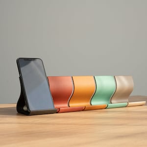 Può includere: Un supporto per telefono nero con uno smartphone sopra. Il supporto è in pelle ed è disponibile in cinque colori diversi: arancione, giallo, verde, beige e marrone.