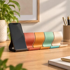 Op de afbeelding: Een rij van vijf gebogen smartphonehouders met een matte afwerking in zwart, terracotta, mosterdgeel, saliegroen en beige. De zwarte houder bevat een smartphone met een donker scherm, terwijl de andere vier houders leeg op een rij staan.