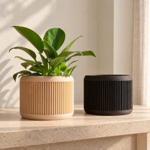 Op de afbeelding: Twee cilindrische plantenpotten met verticale geribbelde texturen. De ene pot is lichtbeige en bevat een groene bladplant met breed, glad blad. De tweede pot is effen zwart en staat leeg naast de beige plantenbak op een vlakke ondergrond.
