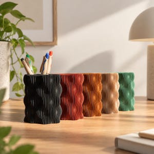 Peut inclure: Une rangée de cinq porte-stylos cylindriques avec une texture ondulée et nervurée verticale. Les supports sont disposés en ligne et sont disponibles en noir, rouge foncé, orange, beige et vert. Plusieurs stylos sont placés dans le porte-stylo noir.